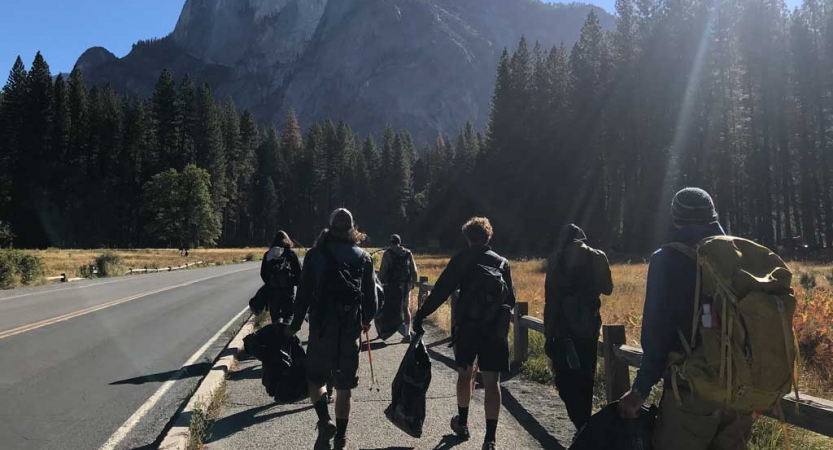 A group of outward bound students carry trash bags as they walk alongside a road during a service project. There are evergreen trees and a mountain in the background. 